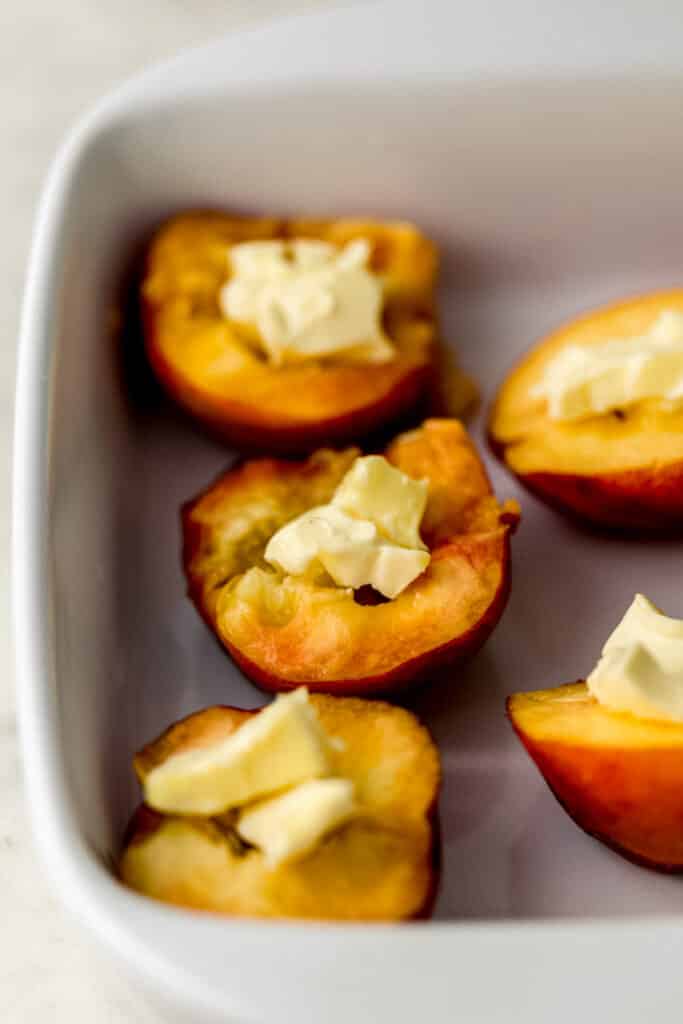 close up side view peaches cut in half with topped with butter in white baking dish 