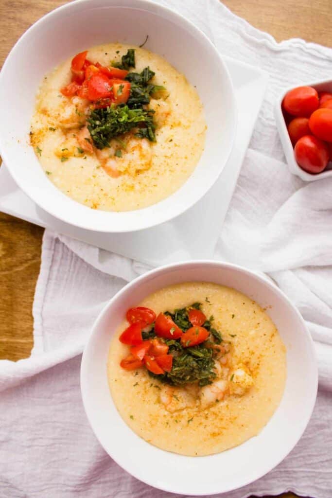 overhead view of two white bowls of shrimp and grits beside small bowl of grape tomatoes 