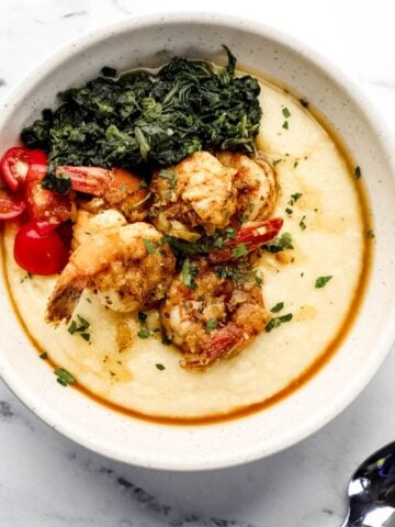 Overhead view of bowl of shrimp and grits by a spoon on marble surface.