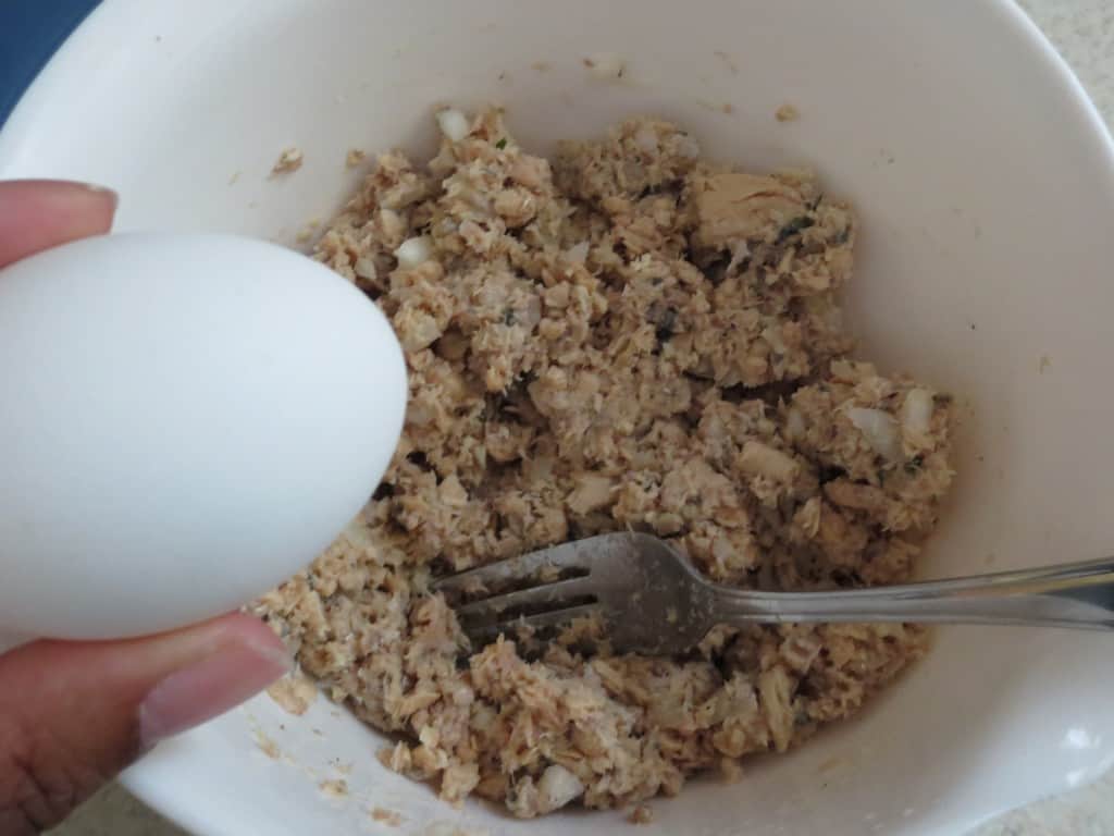 hand adding egg to white mixing bowl with salmon patty mixture with fork