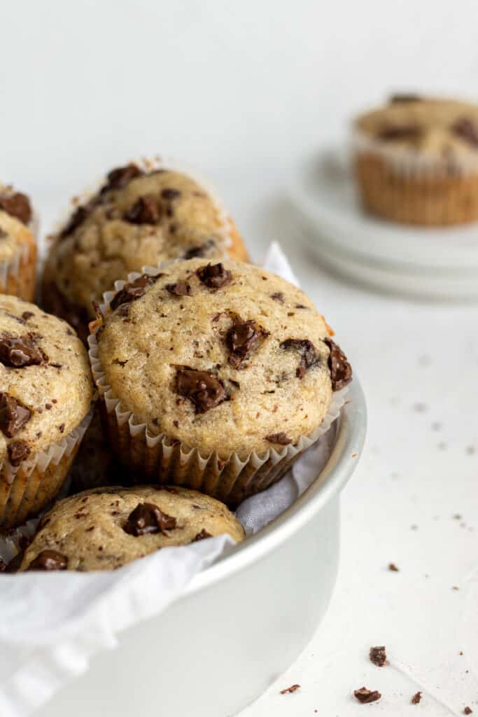 close up side view chocolate banana muffins in white bowl with a single muffin on a plate in the background. 