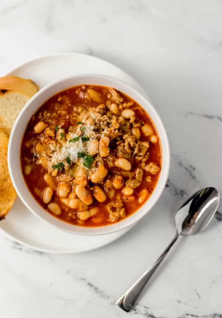 overhead view of white bowl of soup on white plate with bread on it beside a spoon on marble surface 