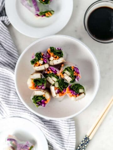 overhead view of finished spring rolls in white plate beside cloth napkin, sauce, plate, and chop sticks