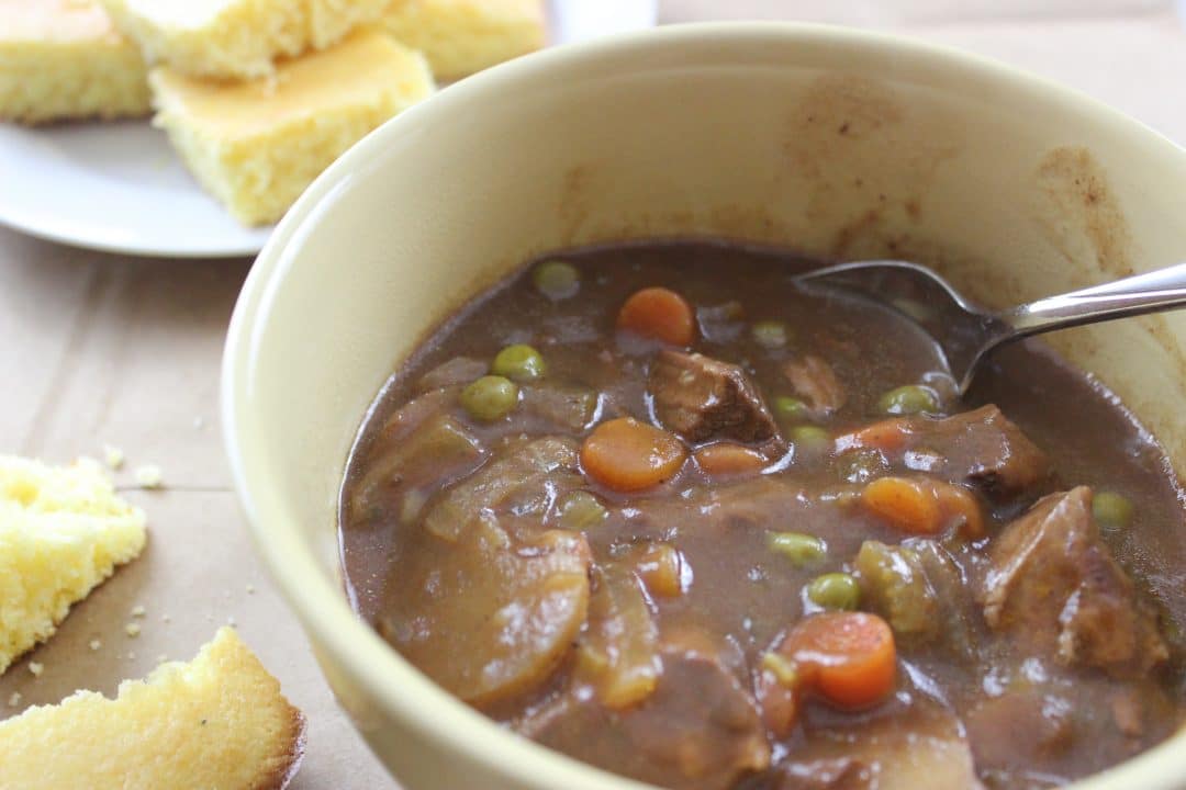 yellow bowl with finished beef stew in it with spoon and cornbread in the background