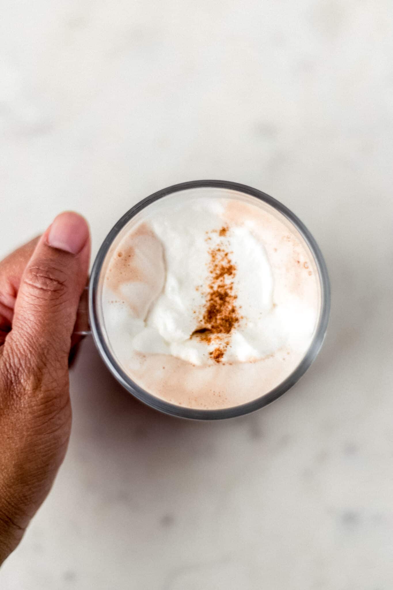 hand on handle of glass cup of hot chocolate topped with whipped topping and a sprinkle of cinnamon