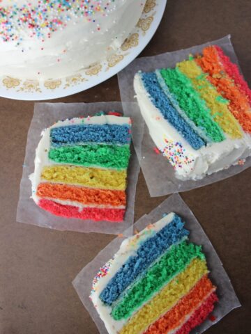 three slices of rainbow cake on wax paper with full cake in the background