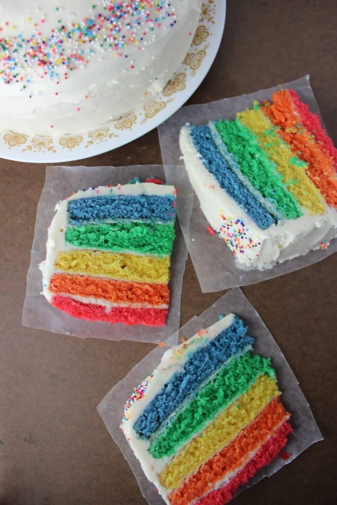 overhead view of three slices of rainbow cake on wax paper with full cake in the background
