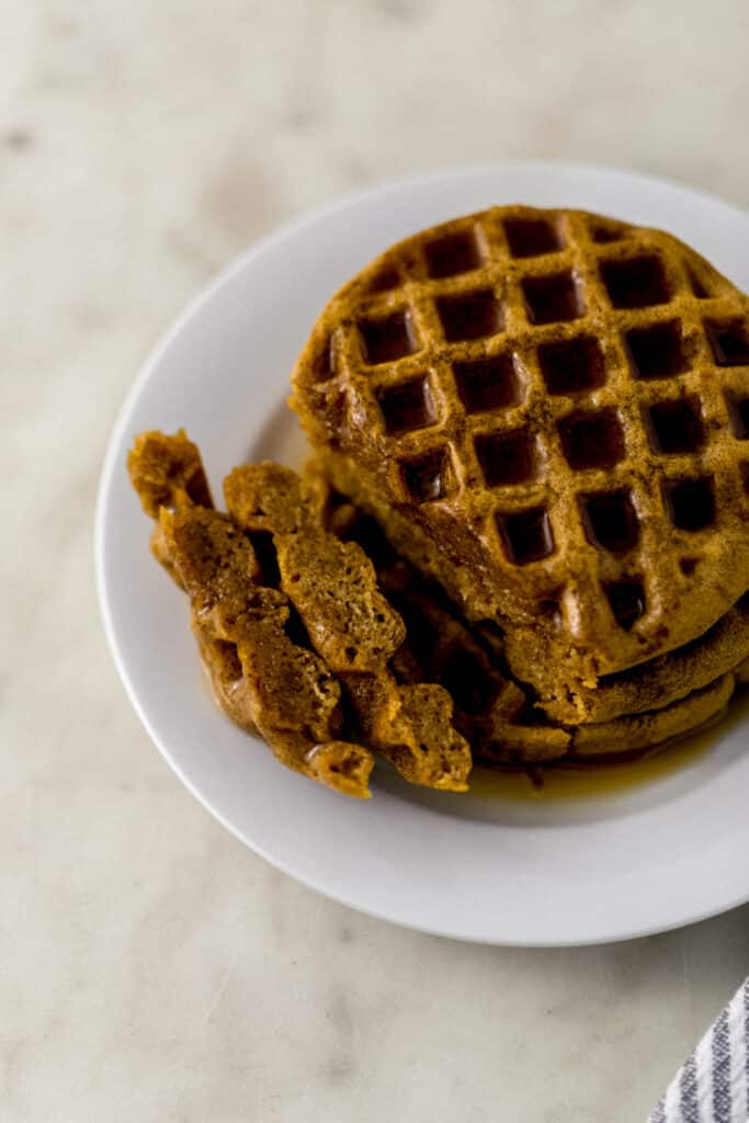 waffles stacked on a white plate topped with maple syrup. 