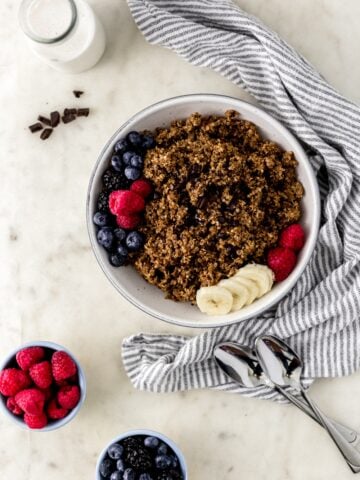 overhead view with chocolate quinoa in white bowl with spoons, cloth napkin, and small bowls with fresh berries.