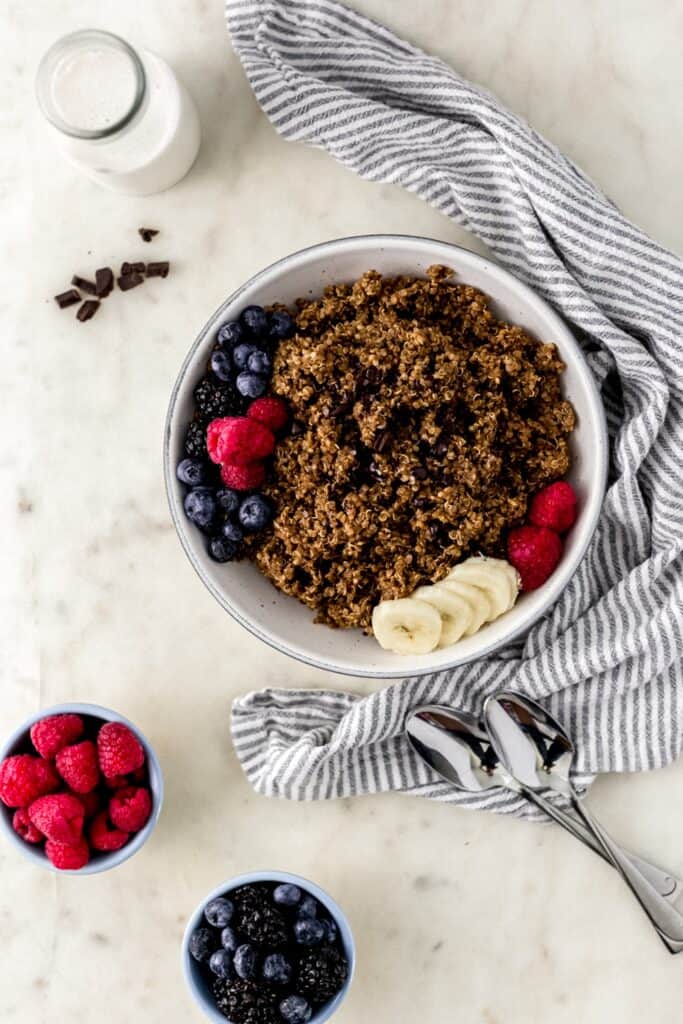 overhead view with chocolate quinoa in white bowl with spoons, cloth napkin, and small bowls with fresh berries. 