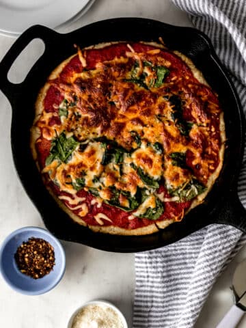 overhead view homemade pizza in cast-iron skillet with red pepper flakes, parmesan cheese, napkin, and pizza cutter