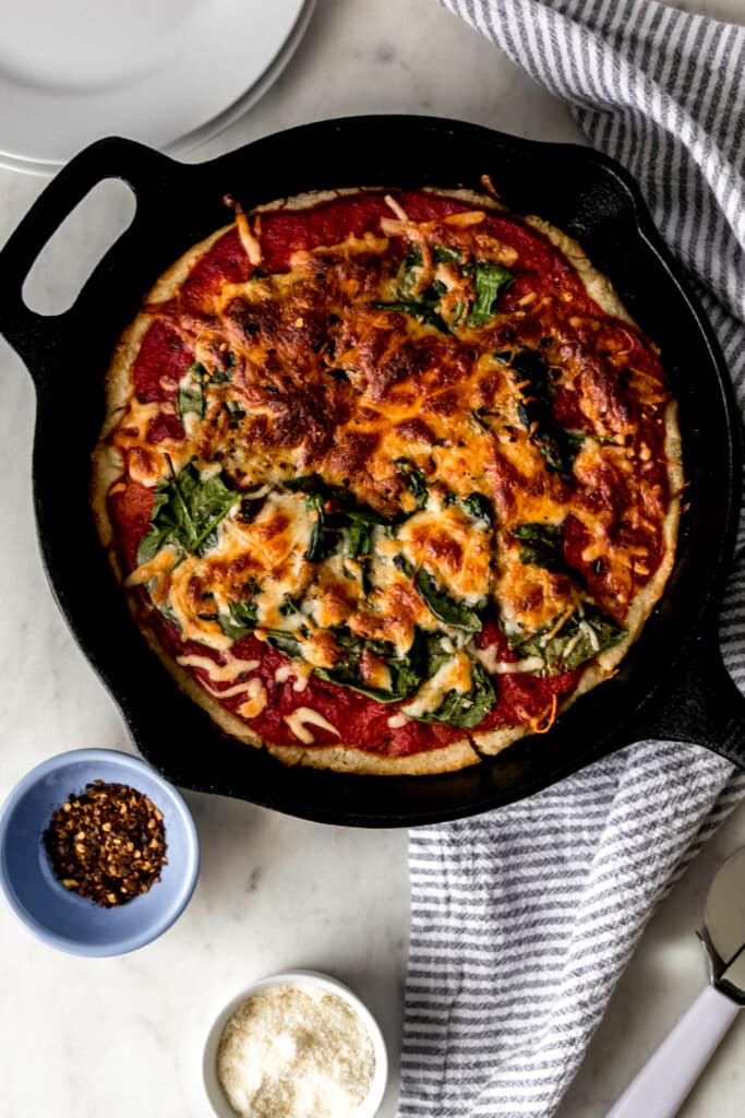 overhead view homemade pizza in cast-iron skillet with red pepper flakes, parmesan cheese, napkin, and pizza cutter 