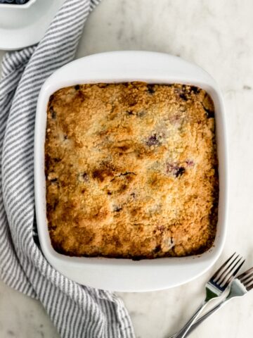 overhead view blueberry cake in white baking dish with white plates, cloth napkin, and forks.