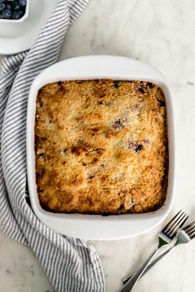 overhead view blueberry cake in white baking dish with white plates, cloth napkin, and forks.