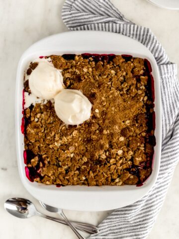overhead view berry crisp dessert in white baking dish with spoons, plates, and napkin