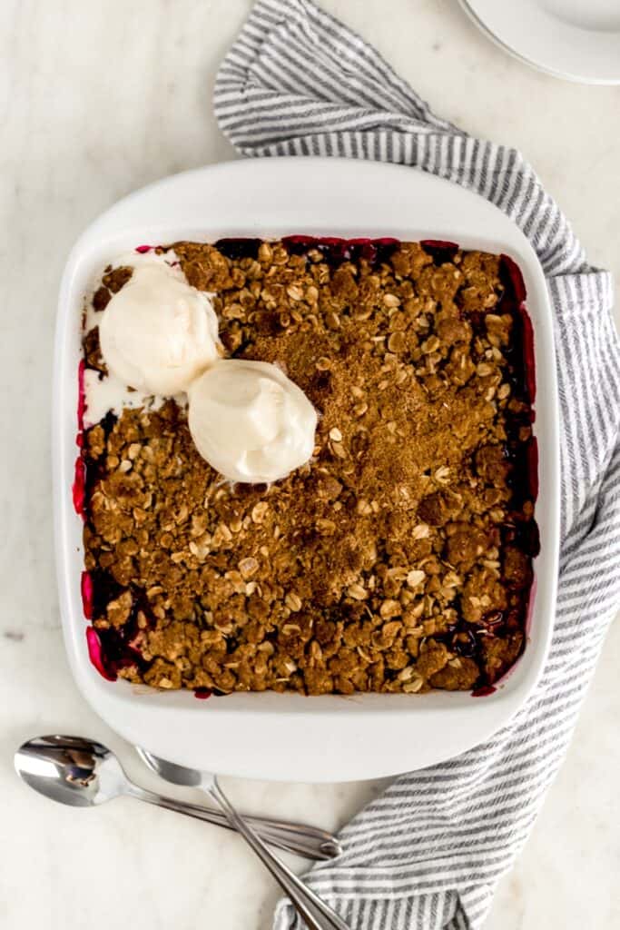 overhead view berry crisp dessert in white baking dish with spoons, plates, and napkin