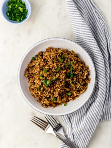 overhead view fried brown rice in white serving bowl with forks, cloth napkin, and small blue bowl with green onion