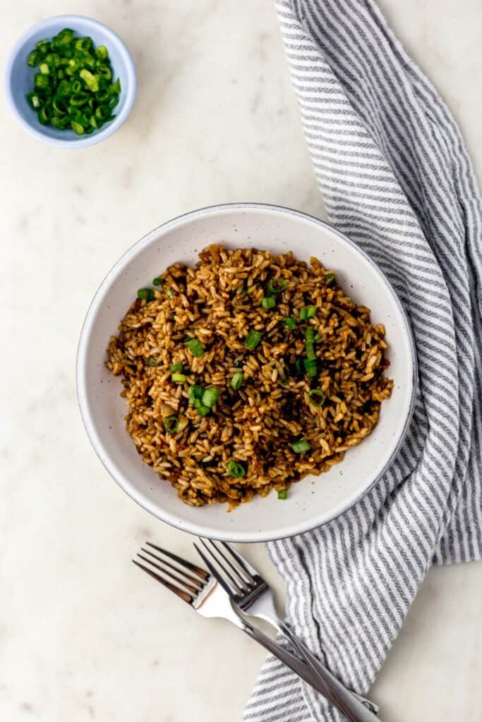 overhead view fried brown rice in white serving bowl with forks, cloth napkin, and small blue bowl with green onion