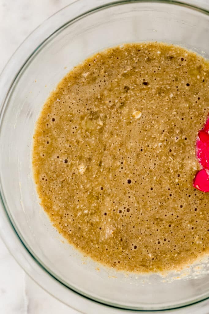 ingredients to make banana pancakes in glass mixing bowl with red rubber spatula