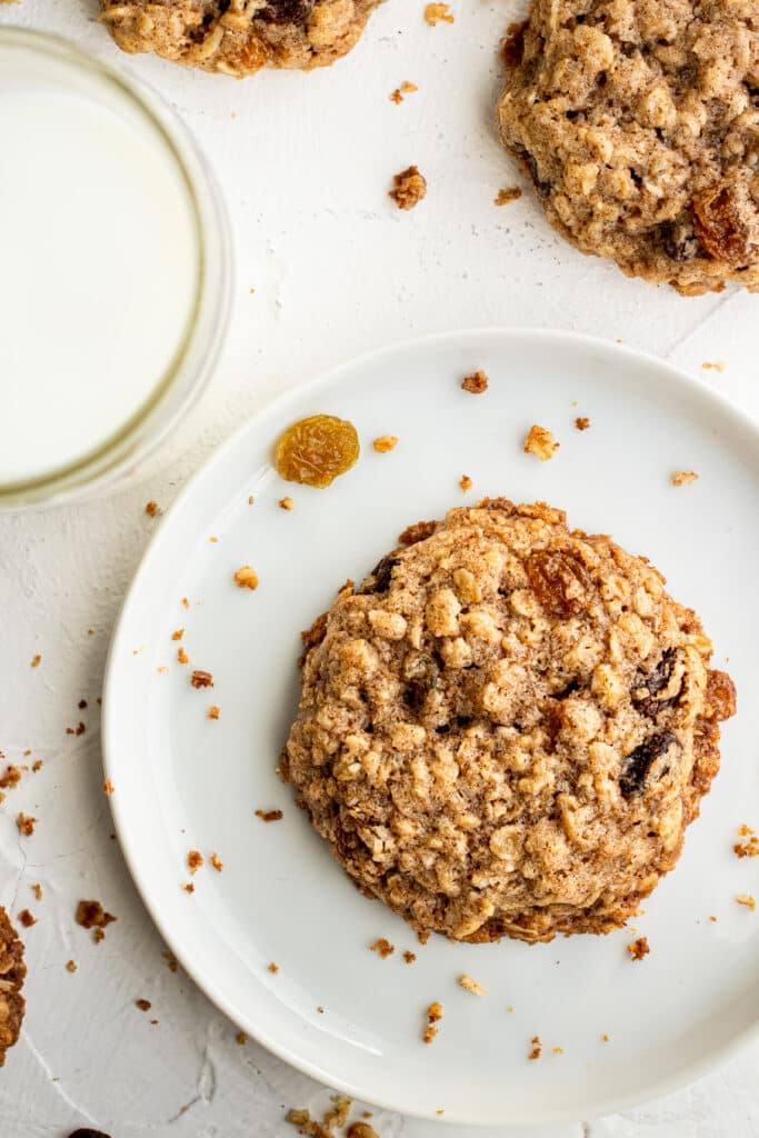 overhead view of single oatmeal raisin cookie on white plate beside a glass of milk.