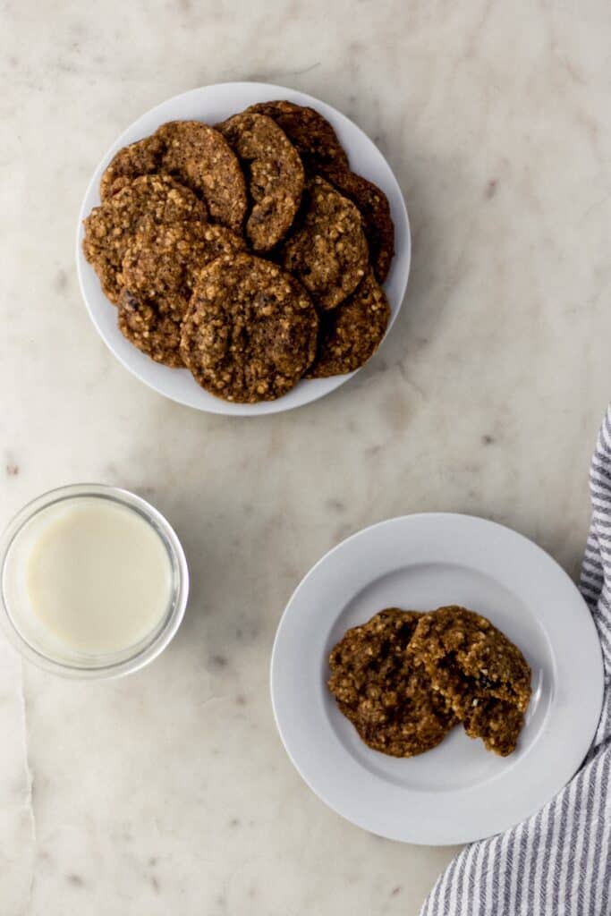 overhead view two white plates with harvest cookies on them next to cloth napkin and glass of milk. 