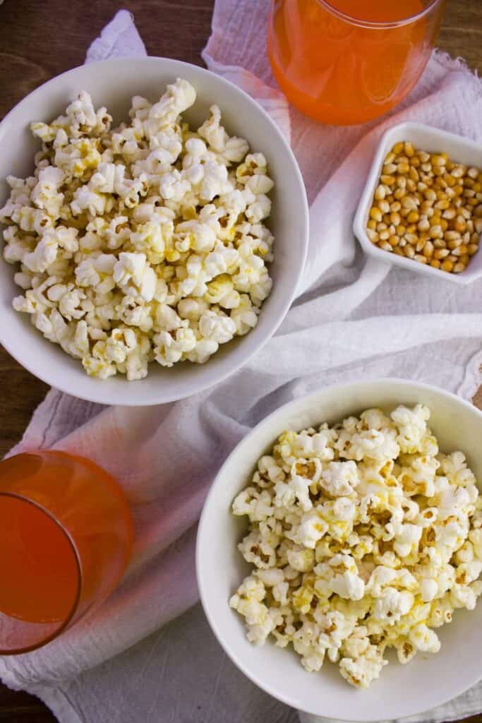 overhead view of popcorn in two white bowls beside cup of orange soda