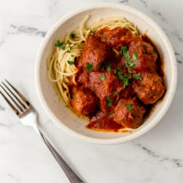 bowl of spaghetti and meatballs beside a fork on marble surface