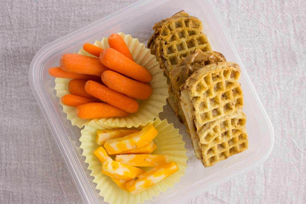 overhead view of peanut butter waffle sandwiches, cheese, and carrots in container 