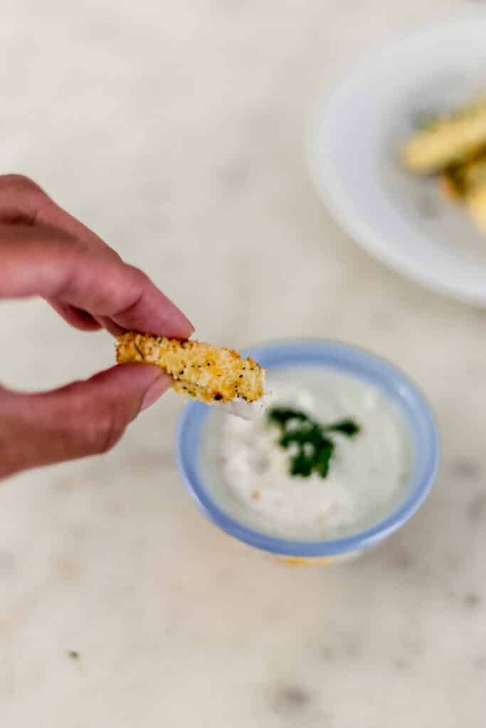 hand holding zucchini fry over container with dipping sauce 