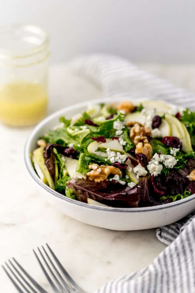 pear and walnut salad in white serving bowl with dressing in small jar and napkin