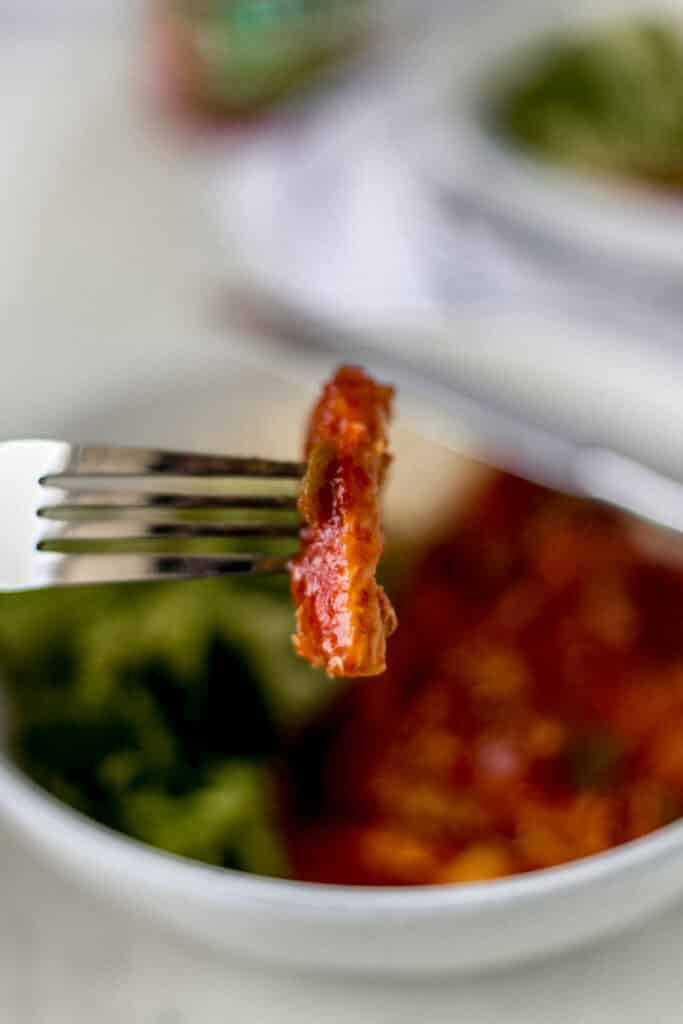 hand holding fork with piece of peachy salsa pork chop on it, plate of food in background