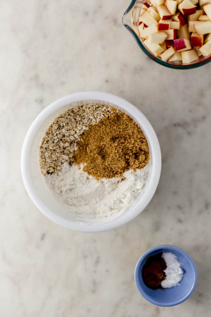 ingredients to make apple crumble bars in small bowls on marble surface. 