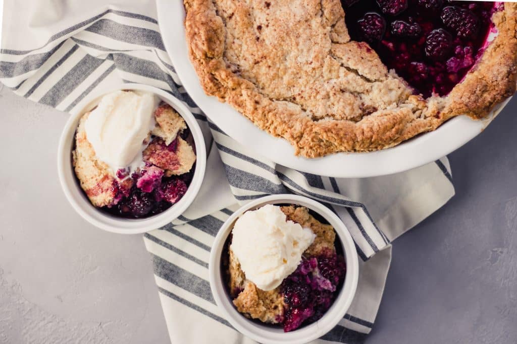 overhead view of finished Blackberry Cobbler in two bowls and the serving dish topped with ice cream 