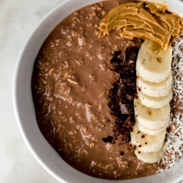 overhead view chocolate mocha overnight oats in white bowl topped with banana slices, coconut, peanut butter, and chocolate