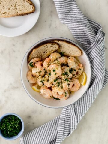 white serving bowl with garlic and butter shrimp next to cloth napkin, white plates, and small bowl with parsley.