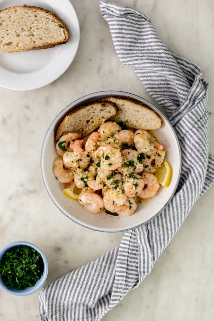 white serving bowl with garlic and butter shrimp next to cloth napkin, white plates, and small bowl with parsley. 