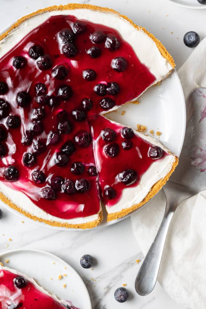 overhead view of blueberry cheesecake with a piece cut out of it on a white plate. 