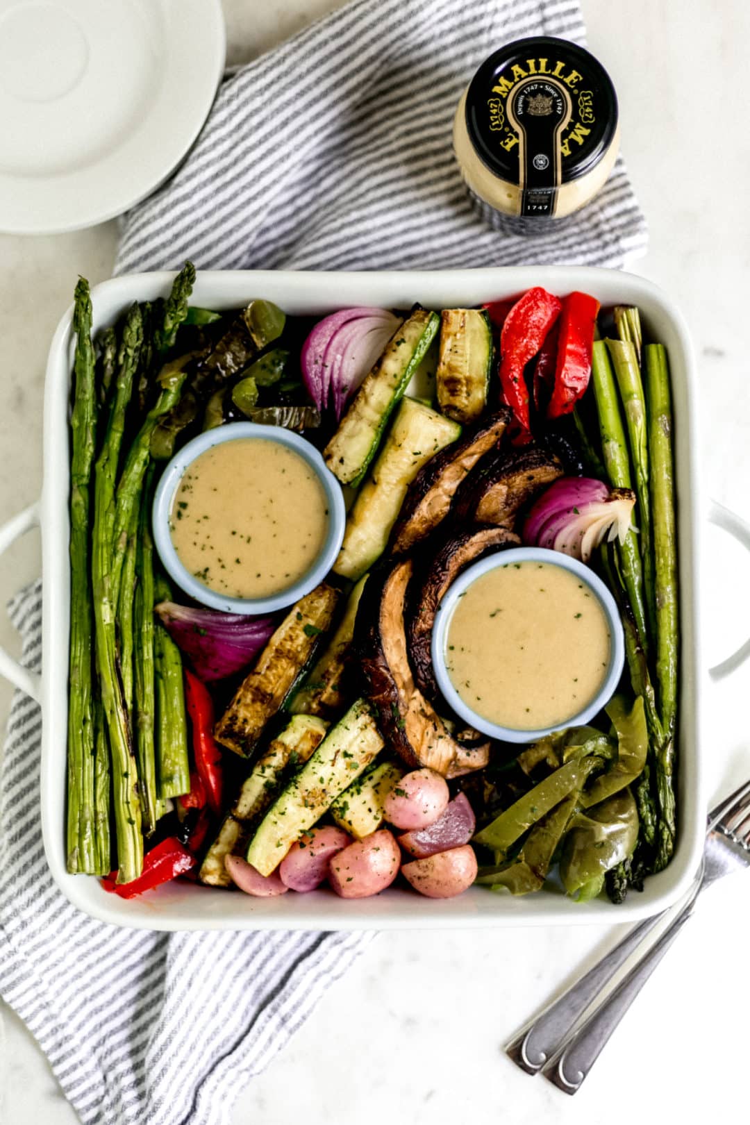 overhead grilled vegetable platter with sauce over napkin with white plates and jar of mustard