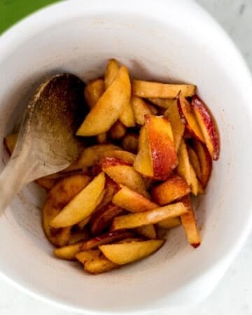Sliced peaches and other ingredients combined in mixing bowl with wooden spoon.