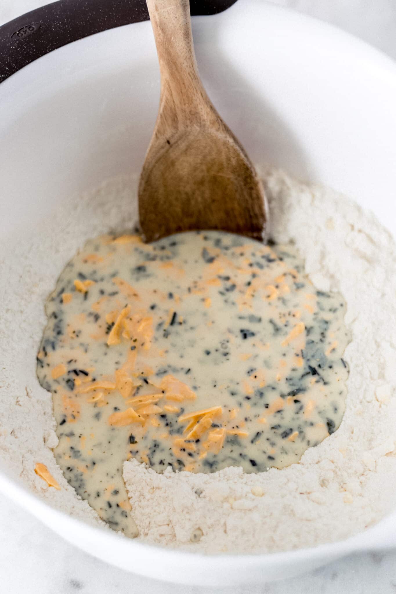 ingredients to make cheddar biscuits in white mixing bowl with wooden spoon