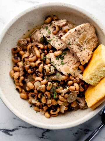 Overhead view of white bowl with black eyed peas, pork chops, and cornbread by a spoon on marble surface.