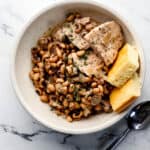 White bowl with black eyed peas, pork, and corn bread in it beside a spoon on marble surface.