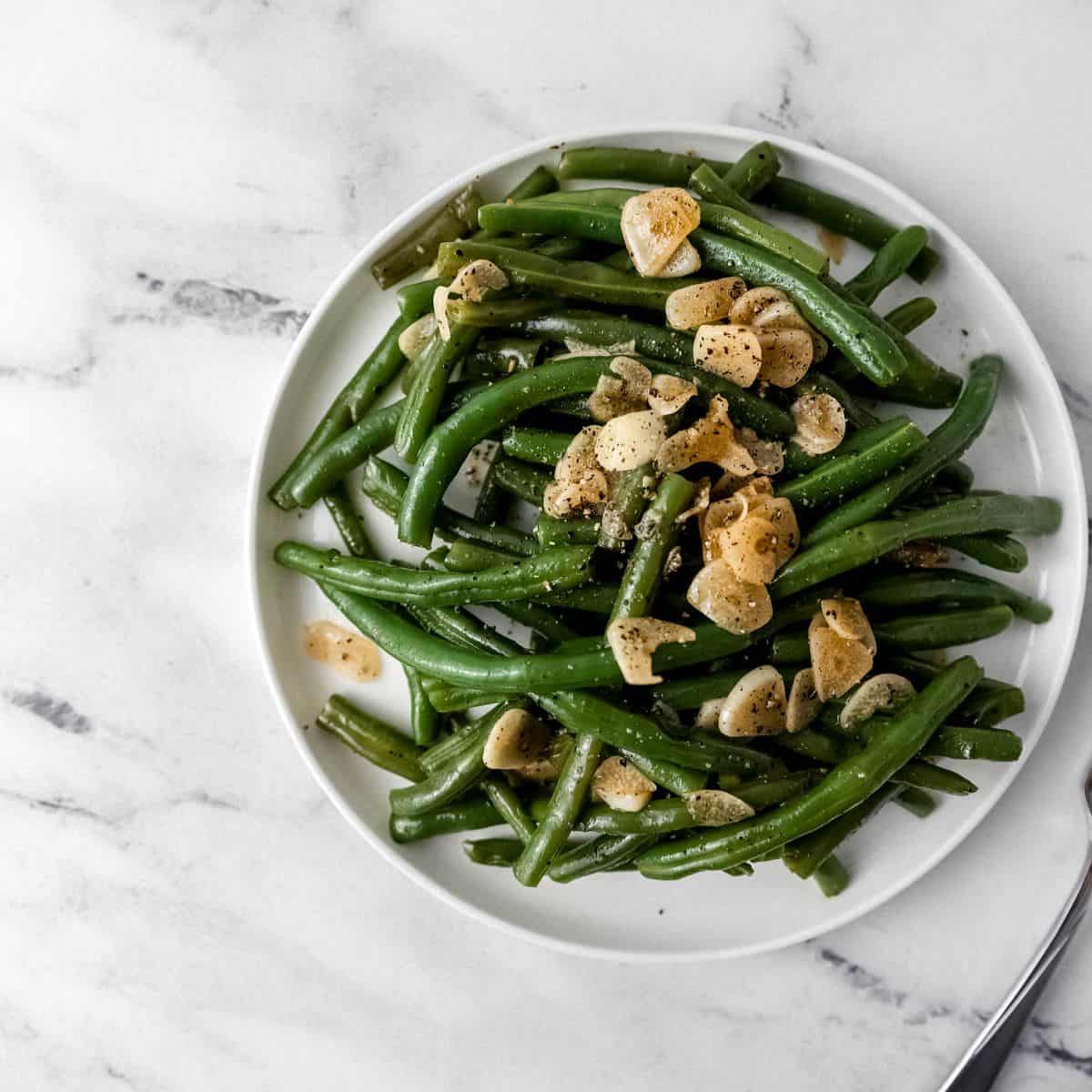 Overhead view of green beans on a white plate by a fork on marble surface.
