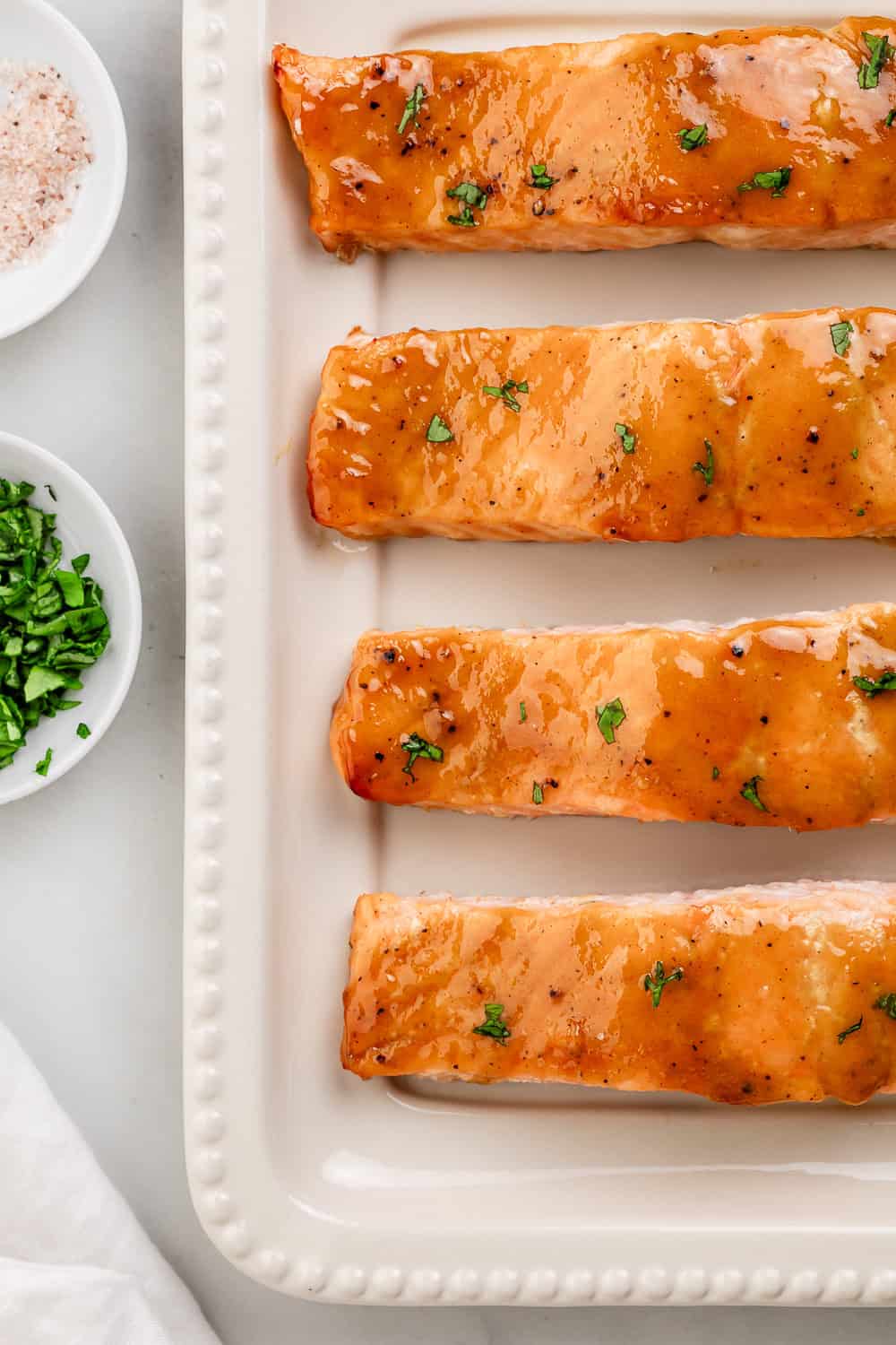 Overhead view of finished salmon fillets on white platter by small bowls of salt and parsley.