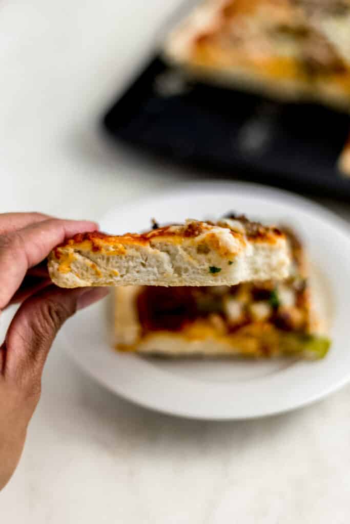 hand holding piece of supreme pizza over white plate with another piece in the background 