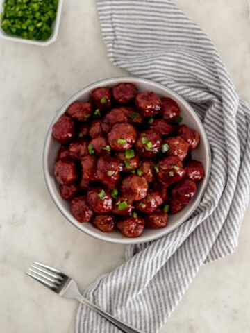 sweet and spicy meatballs in a white serving bowl with fork and napkin