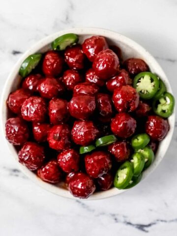 Overhead view of finished meatballs and slice jalapenos in white bowl on marble surface.