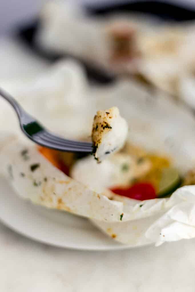 hand holding fork with piece of cod over parchment packet on white plate