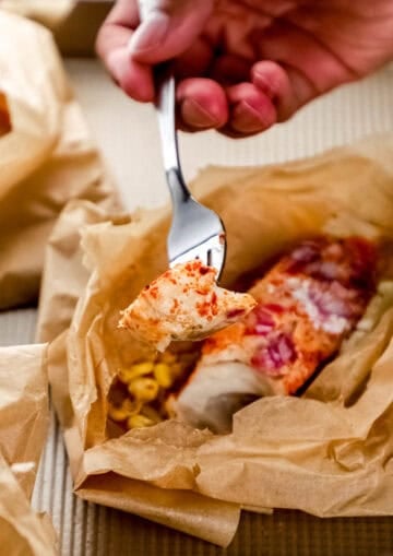 Hand holding fork with fish on it over the packet on sheet pan.