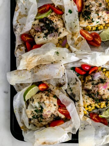 baked cod packets in parchment paper on baking sheet. Packets have fish, corn, red onion, tomatoes, and lime slices.